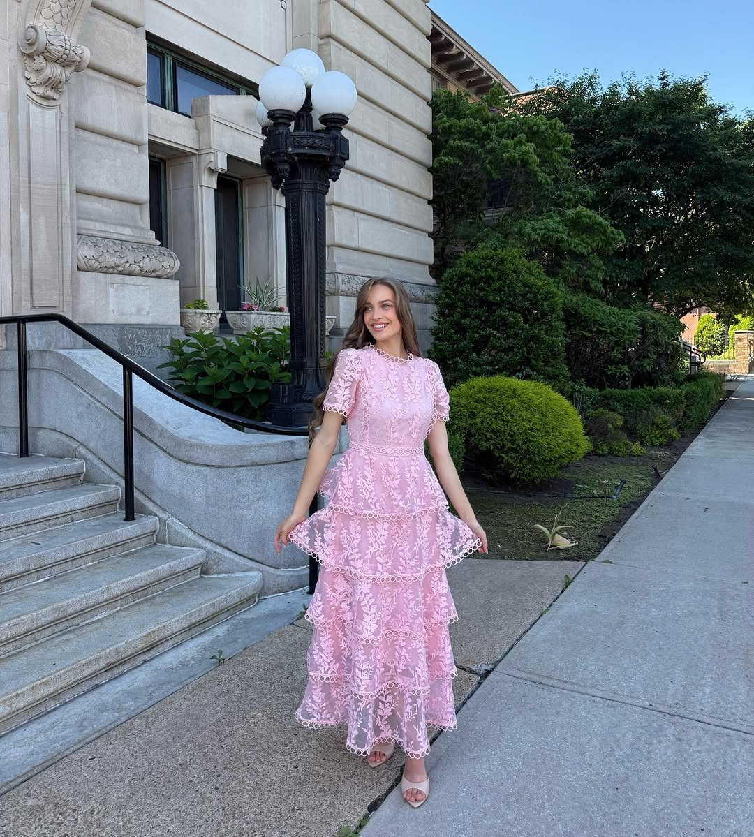 Woman wearing a tiered pink lace midi dress standing on a city sidewalk.