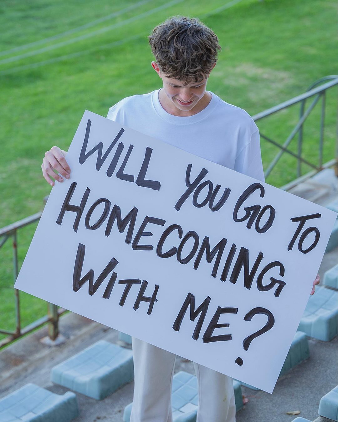 Young man holding a white poster asking someone to homecoming on bleachers