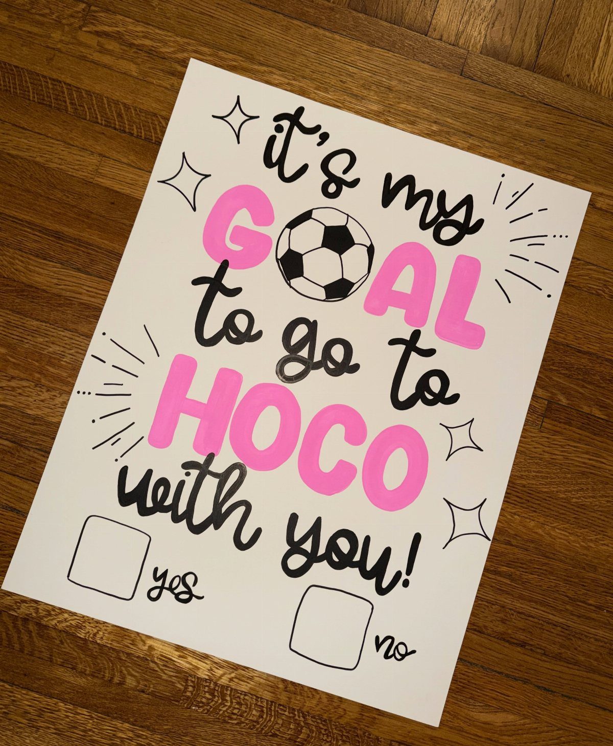 White homecoming proposal poster with soccer ball and pink lettering on wooden floor